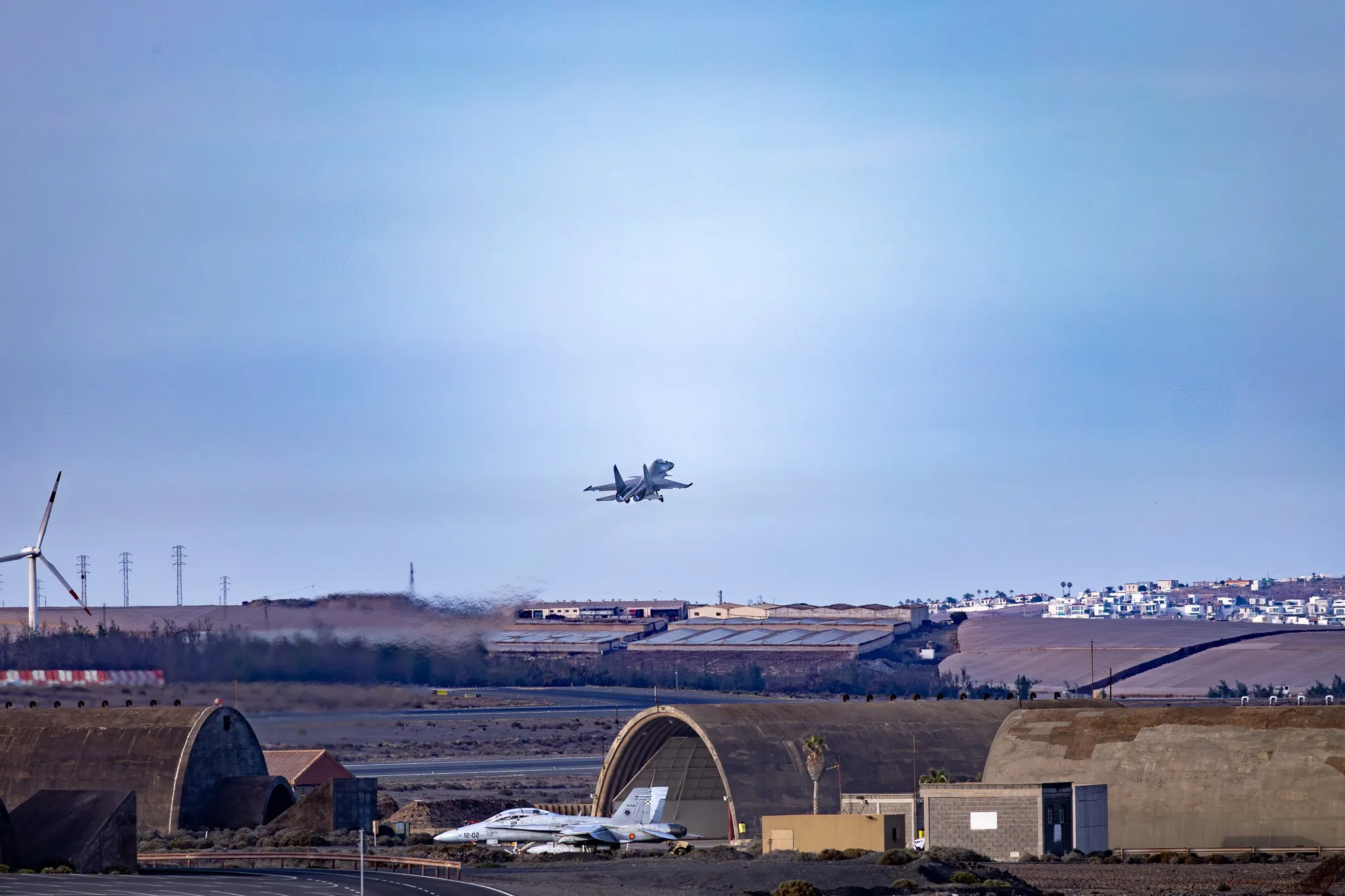 Ein Kampfjet vom Typ F/A-18 Hornet im Landeanflug im Tiefflug über halbrunde Splitterschutz-Hangars, im Vordergrund eine baugleiche Maschine am Boden, im Hintergrund ein Windrad und eine Stadt am Hang, klarer blauer Himmel.