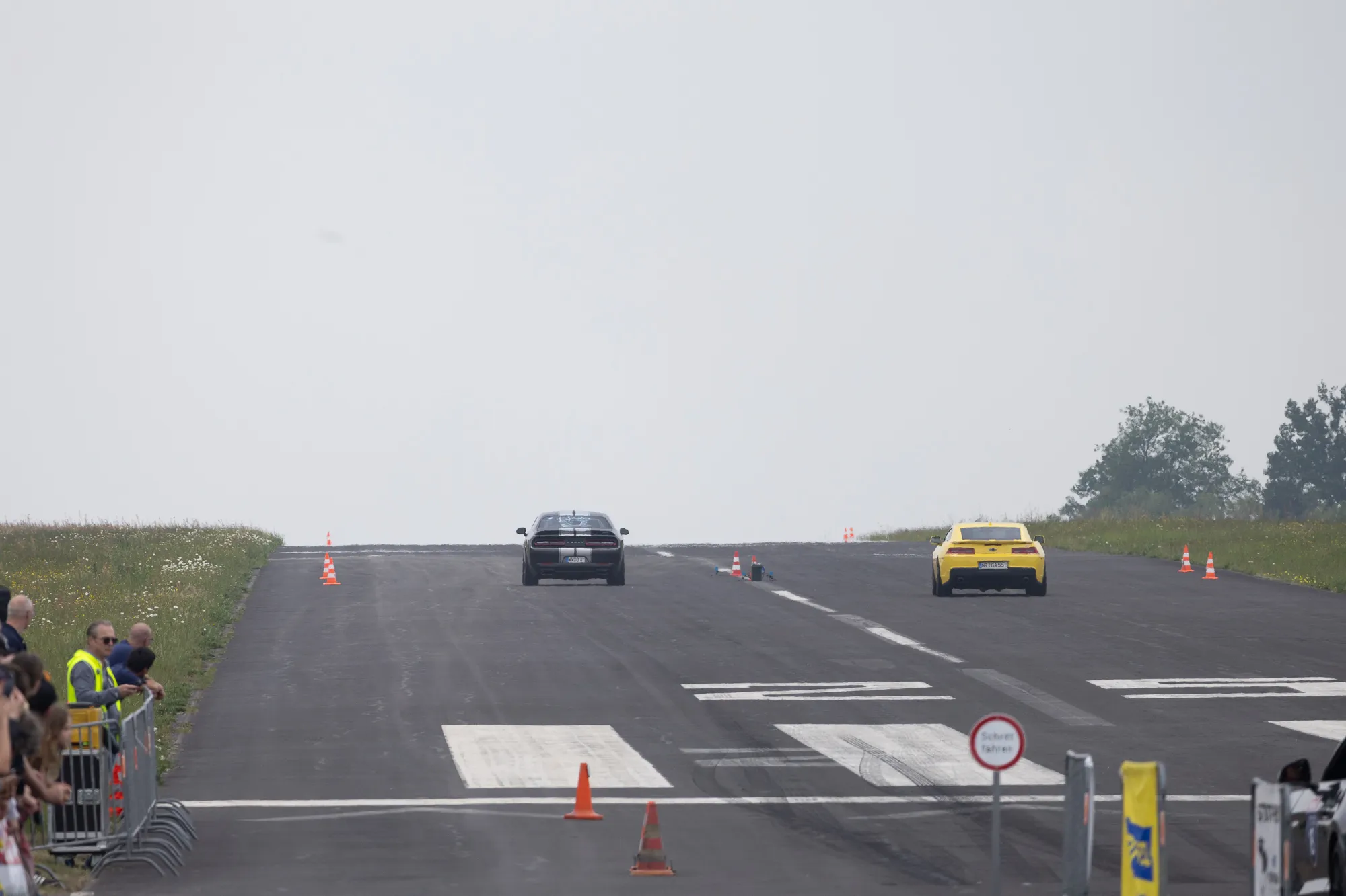 Blick die Startbahn des Flugplatzes Meinerzhagen hinunter: Ein dunkler Dodge Challenger und ein gelber Chevrolet Camaro rollen im Nebel Seite an Seite über die markierte Rennstrecke, links Zuschauer hinter Absperrgittern.