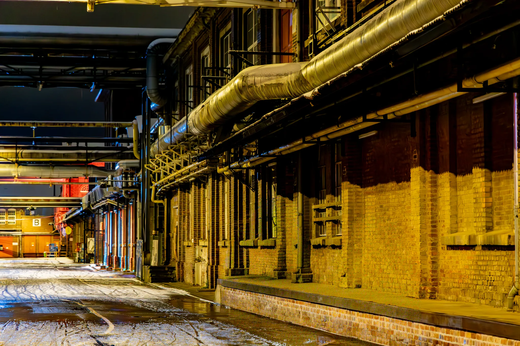 Schmaler Werksweg zwischen Backsteingebäuden auf dem Zanders Gelände bei Nacht, darüber ein silbern glänzendes Rohrleitungssystem, der nasse Boden spiegelt warmes Kunstlicht.