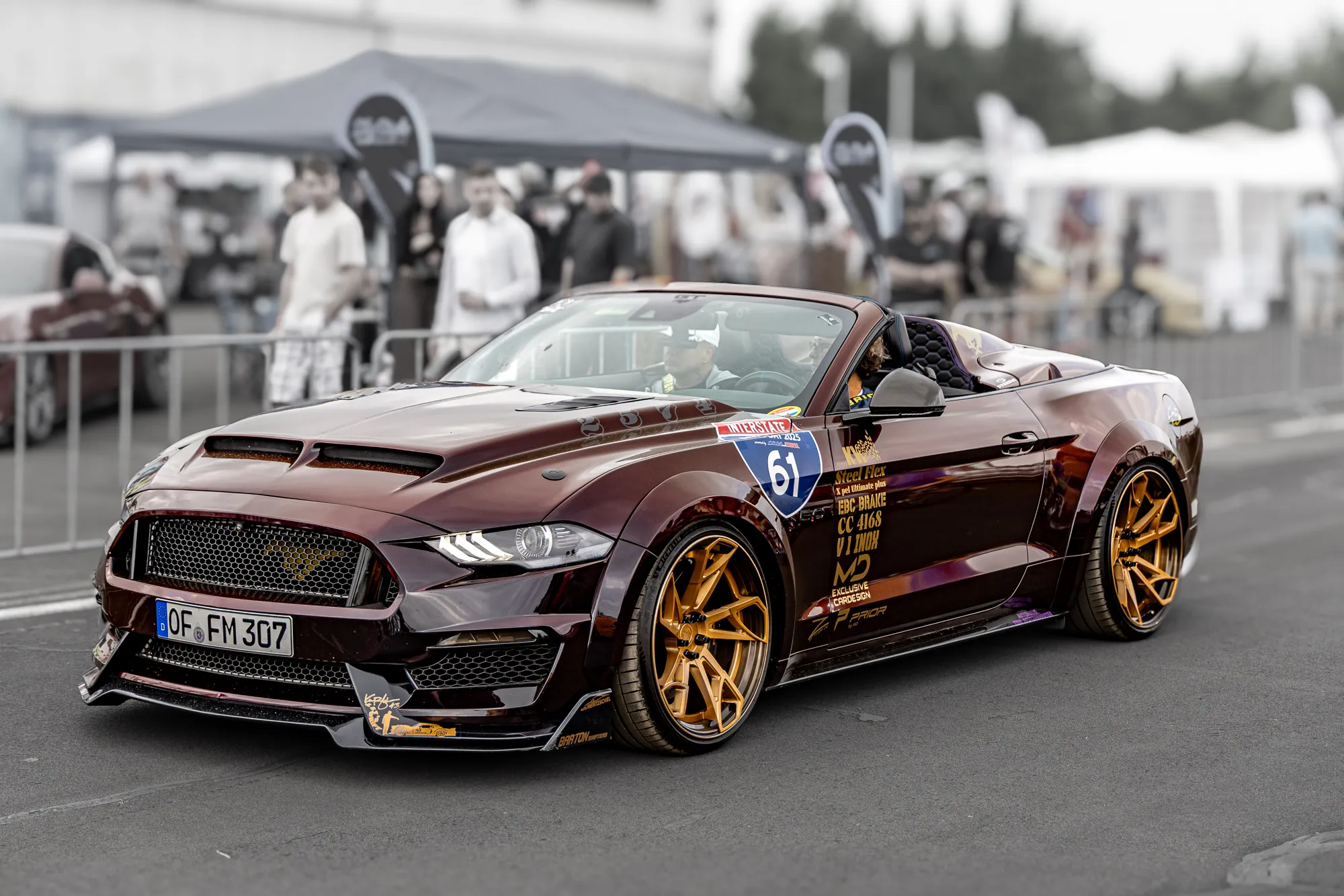 Dunkelroter, breit ausgestellter Ford Mustang Cabrio mit goldenen Felgen und Startnummer 61 rollt an einer Zuschauermenge unter Pavillons vorbei.