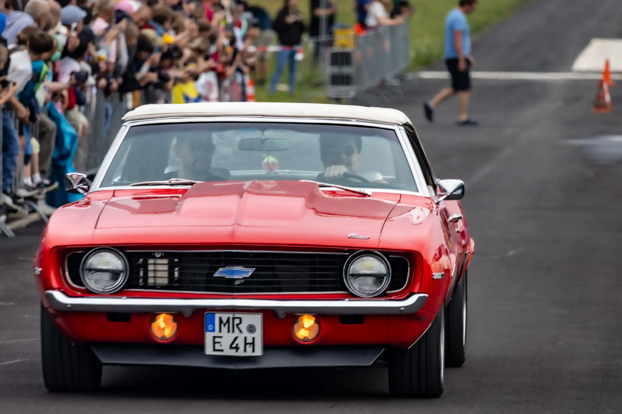 Roter Chevrolet Camaro Cabrio mit weißem Verdeck fährt frontal auf die Kamera zu, links jubelnde Zuschauer hinter Absperrgittern, im Hintergrund ein Ordner und Pylonen.