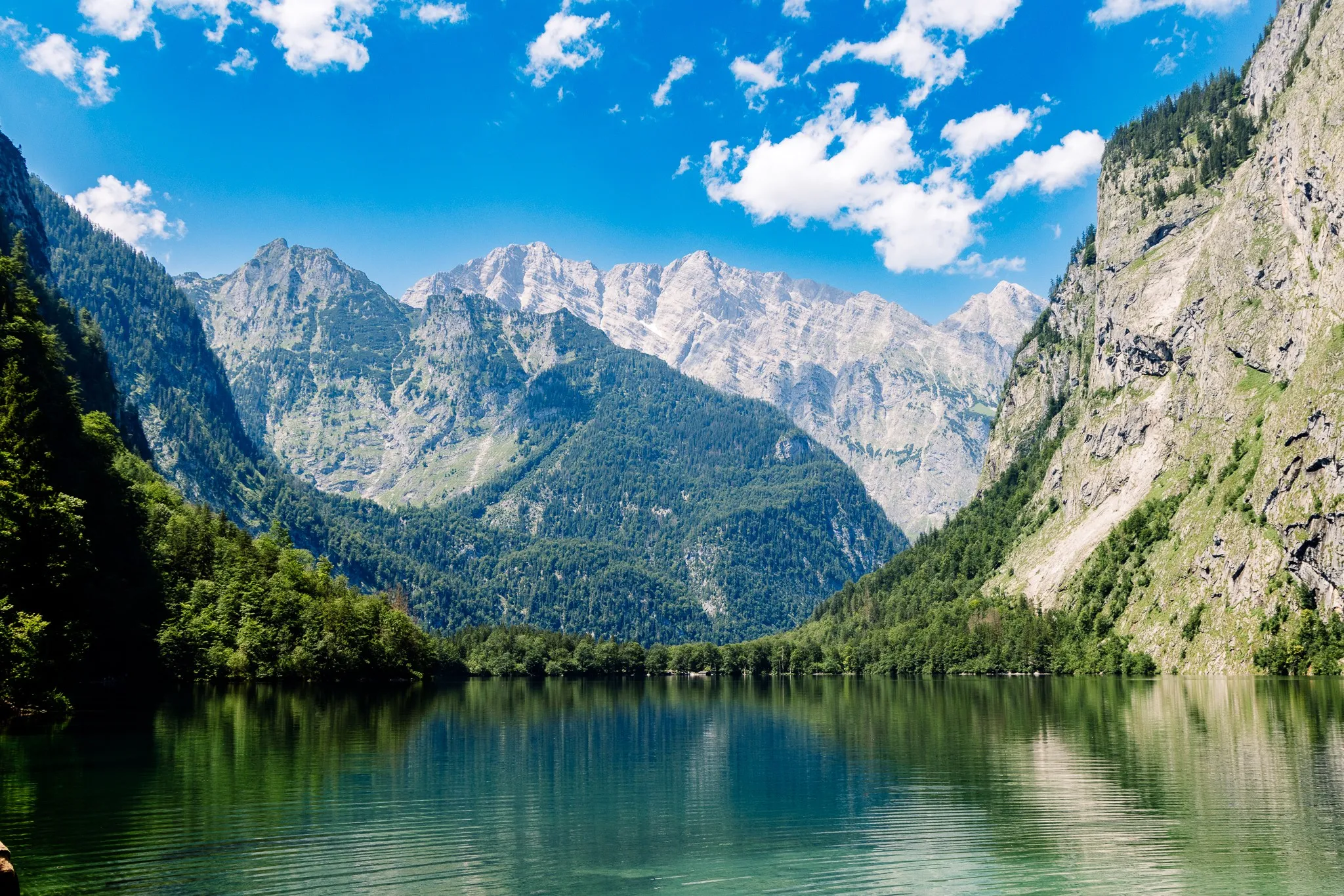 Ein alpiner Bergsee liegt zwischen bewaldeten Hängen und schroffen Felsgipfeln, gerahmt von einer massiven Felswand unter klarem, sonnigem Himmel.