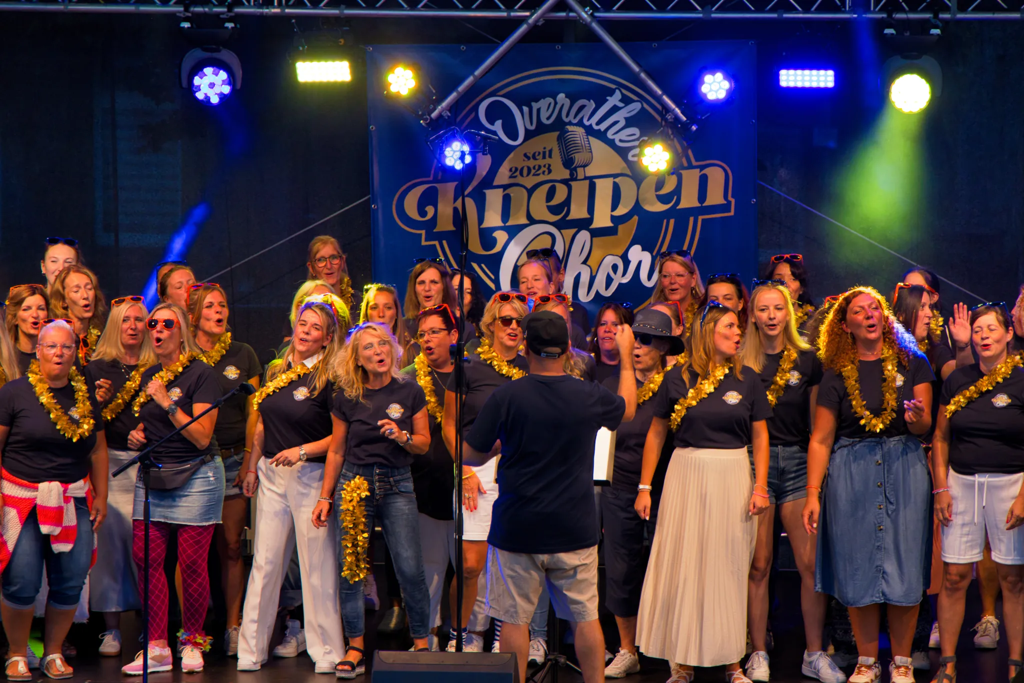 Nahaufnahme des singenden Chors mit goldenen Blumenketten, im Vordergrund ein Chorleiter mit Rücken zur Kamera vor dem Bühnenbanner „Kneipenchor“.