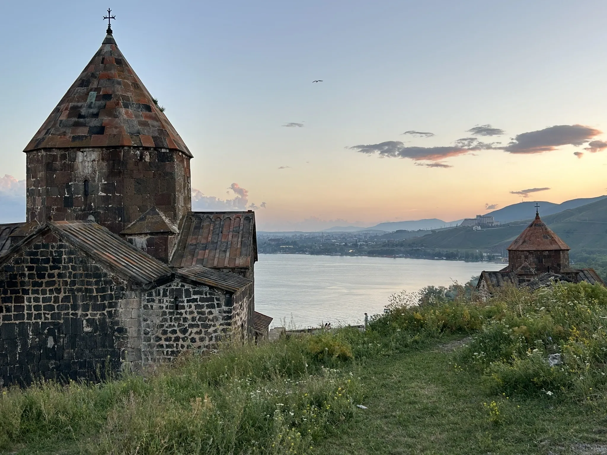 Eine historische steinerne Kirche steht am Seeufer, im Vordergrund Wiese mit gelben Blumen, während der Abendhimmel in warmes Rosa getaucht ist.