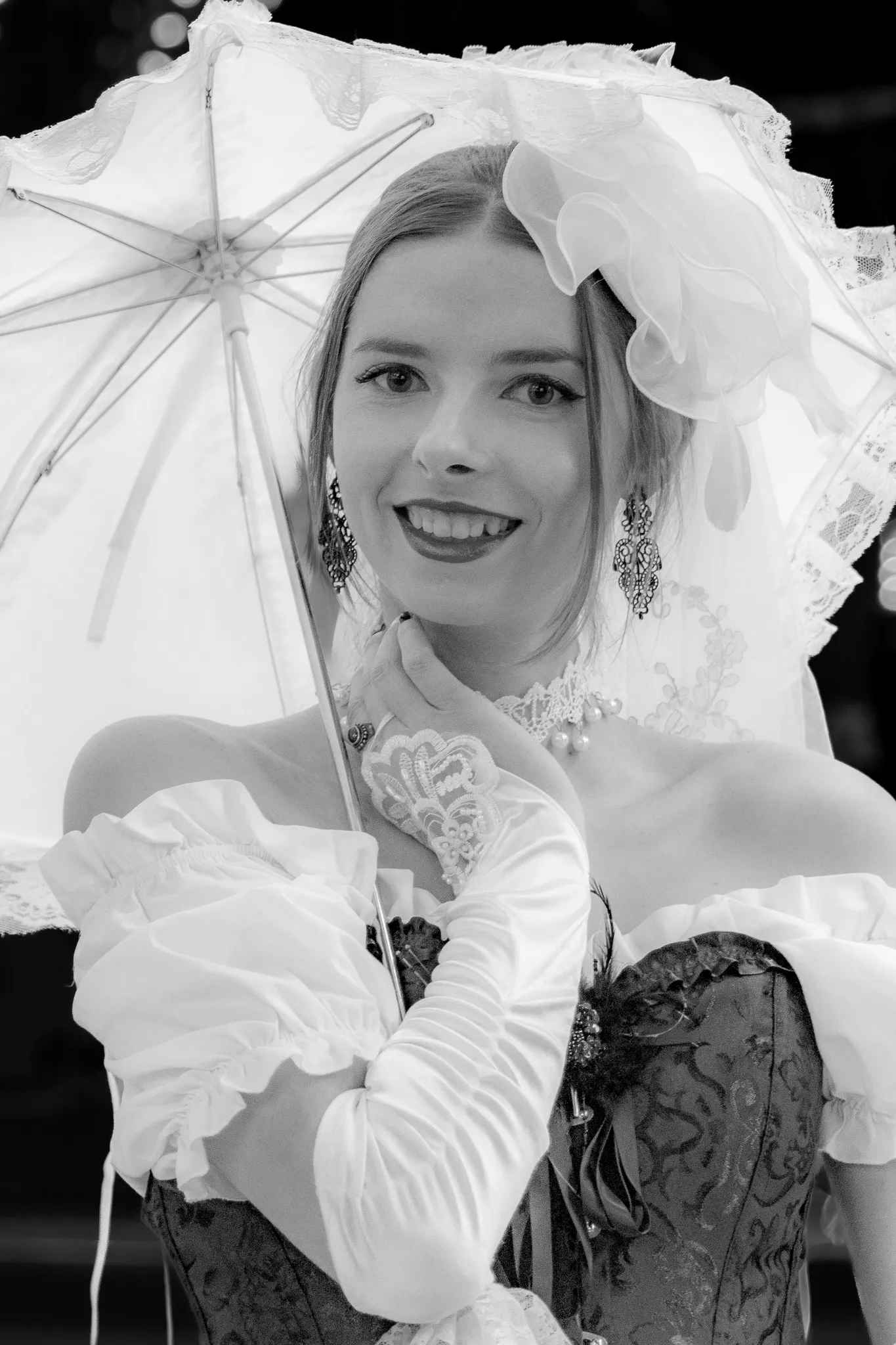 Schwarz-Weiß-Porträt einer jungen Frau unter einem weißen Spitzenschirm, mit Blumenschmuck im Haar, Perlencollier und schwarzem Mieder-Kleid, lachend in die Kamera blickend.