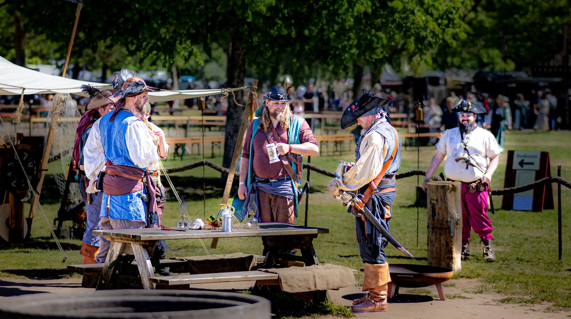 Gruppe von Piraten-Cosplayern in bunten Wamsen, Dreispitzhüten und mit Säbeln, versammelt an einem rustikalen Holztisch mit Flaschen im Freien.