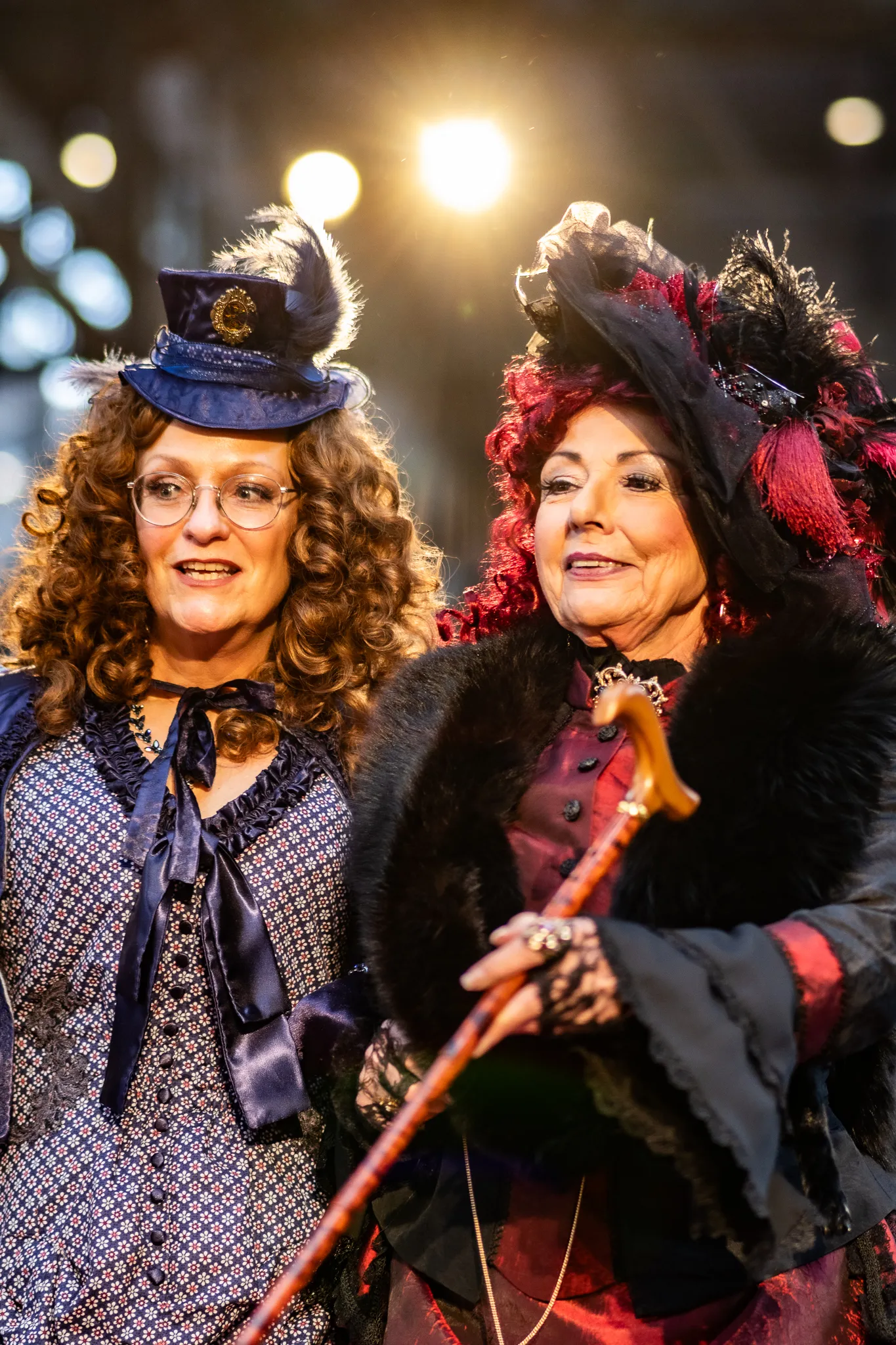 Zwei Frauen in viktorianisch inspirierten Steampunk-Kostümen nebeneinander, eine mit kleinem blauem Zylinder und Lockenperücke, die andere mit Pelzbesatz und Federhut, dahinter Lichter des Jahrmarkts.