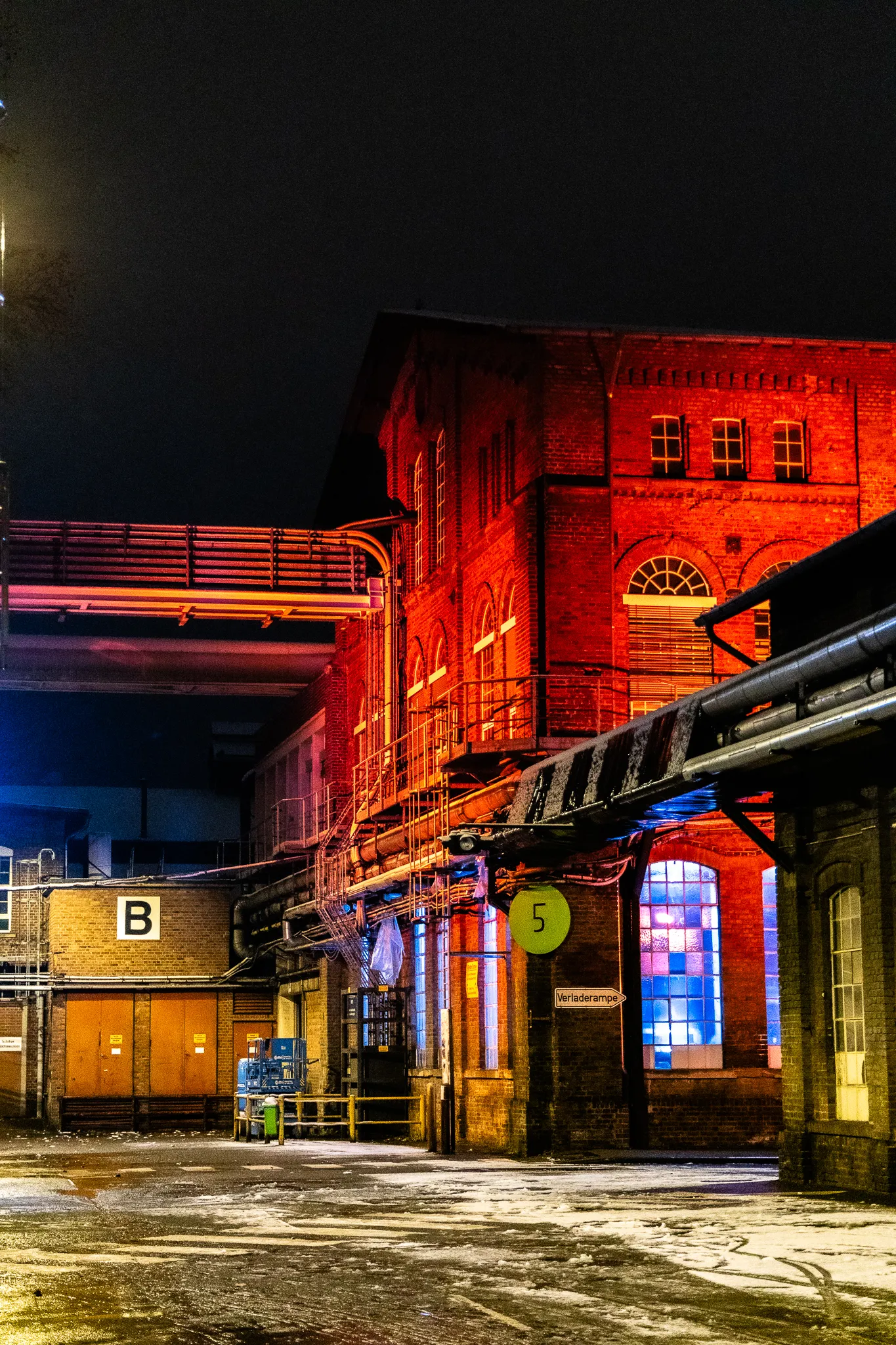 Weiter Blick auf das rot illuminierte Zanders-Backsteingebäude mit Verladerampe, Rohrbrücke und Hoftor „B“ bei Nacht.
