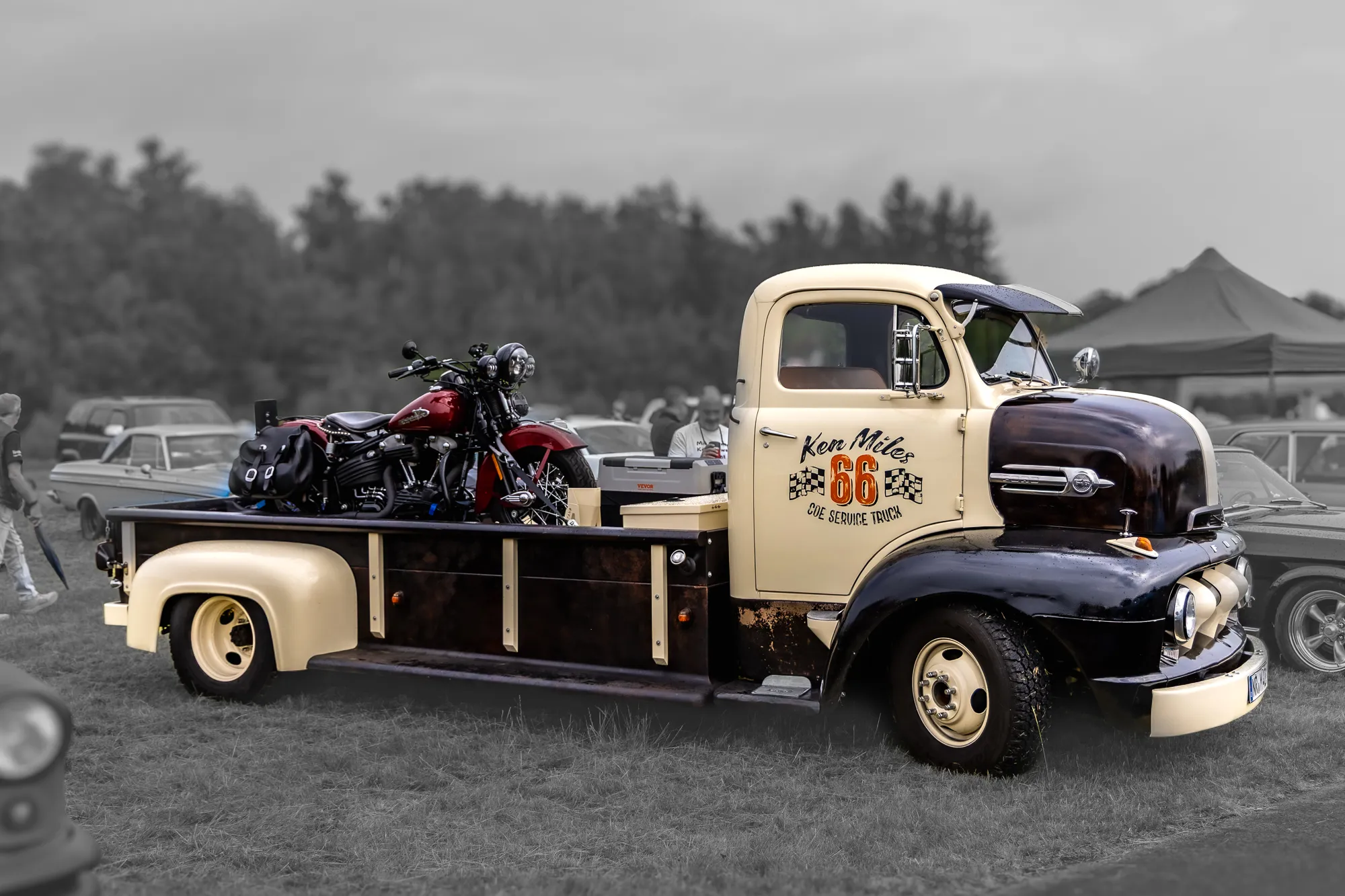 Umgebauter Ford COE-Servicetruck in Creme und Rostbraun mit Schriftzug „Ken Miles 66 COE Service Truck“, auf der Ladefläche eine dunkelrote Harley-Davidson-Motorrad.