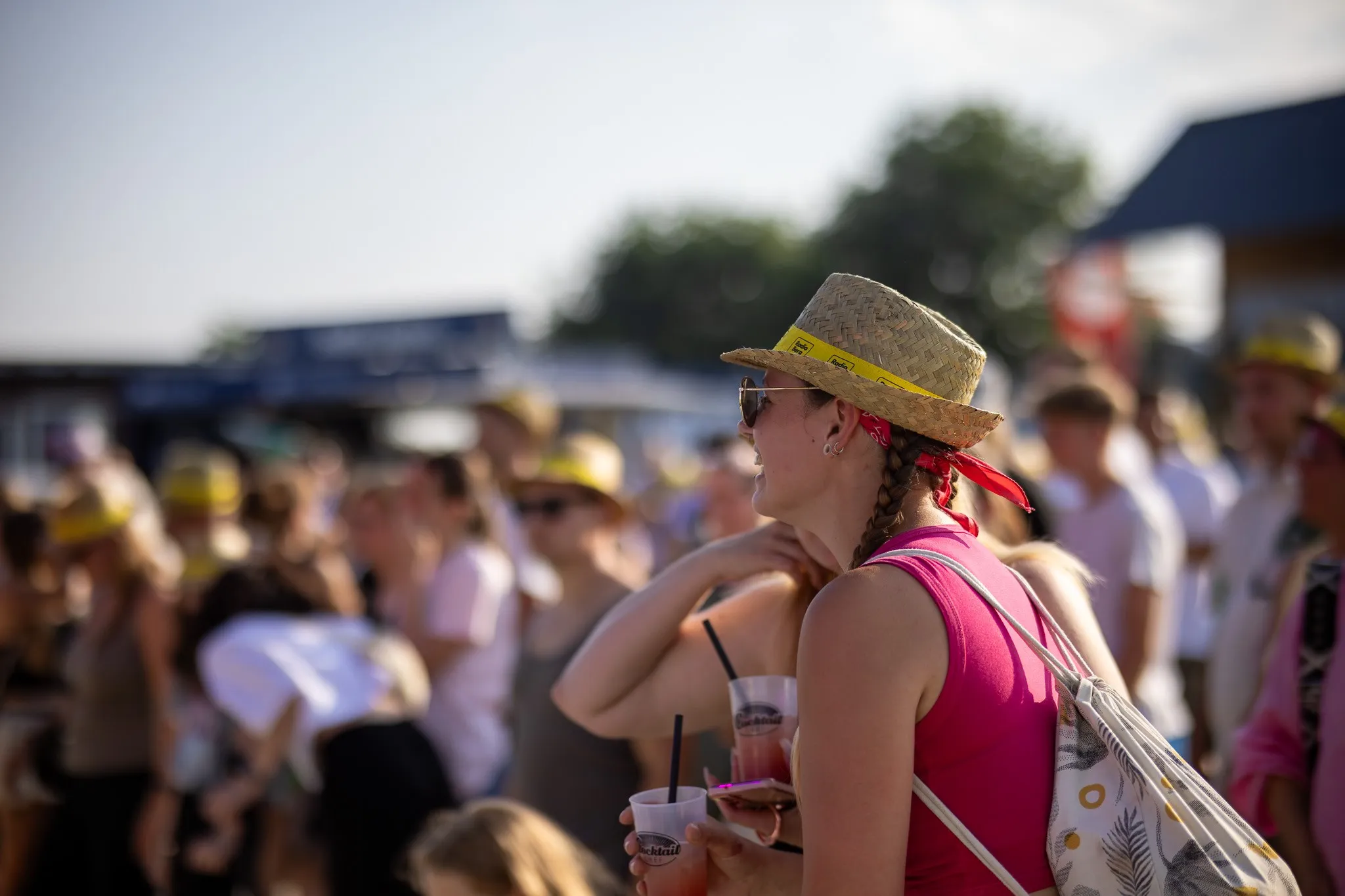 Junge Frau mit Strohhut und zwei Getränkebechern in der Hand steht im Profil inmitten der Festivalmenge, im Hintergrund verschwommene Zelte.