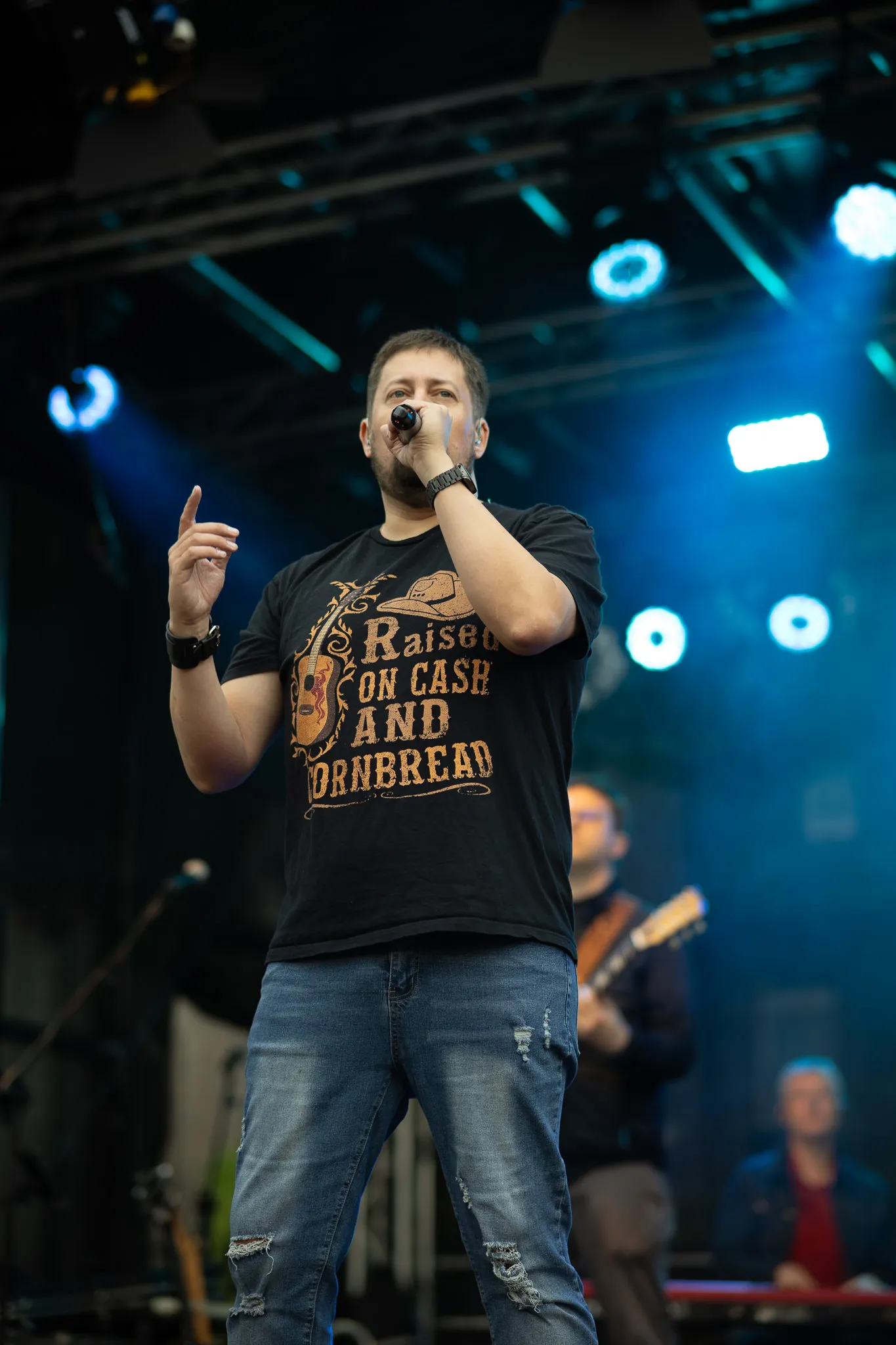 Ein Sänger mit Bart hält das Mikrofon nah an den Mund und hebt den Zeigefinger, sein schwarzes T-Shirt trägt den Aufdruck „Raised on Cash and Cornbread”, im unscharfen Hintergrund sind ein Gitarrist und ein weiteres Bandmitglied zu erkennen.