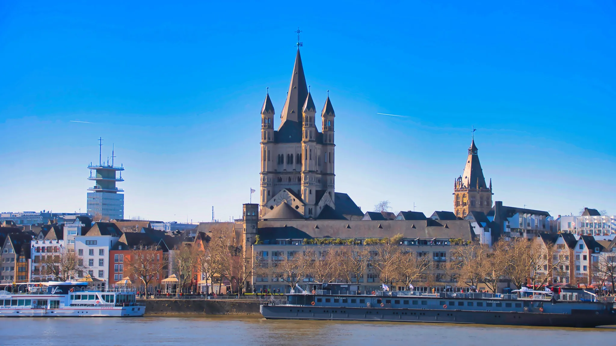Tagesansicht von Köln mit Kölner Dom und Römerturm über dem Rhein, im Vordergrund zwei Passagierschiffe, unter kräftig blauem Himmel.
