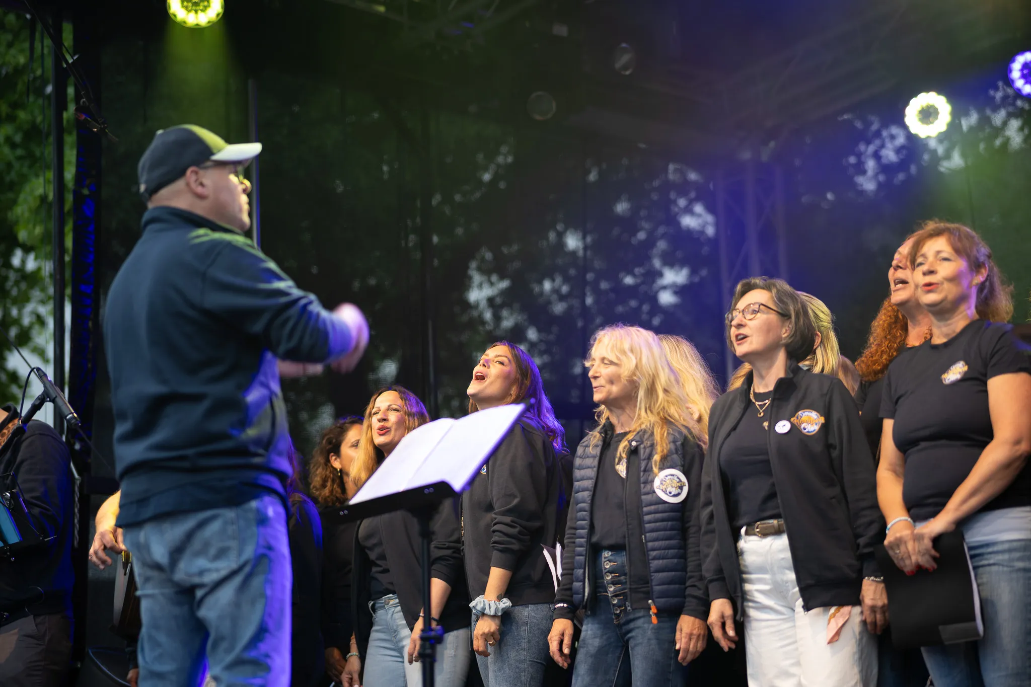 Ein Chorleiter mit Kappe dirigiert von der Seite eine Gruppe singender Frauen in schwarzen Oberteilen, im Hintergrund sind Bäume und Bühnenbeleuchtung zu erkennen.