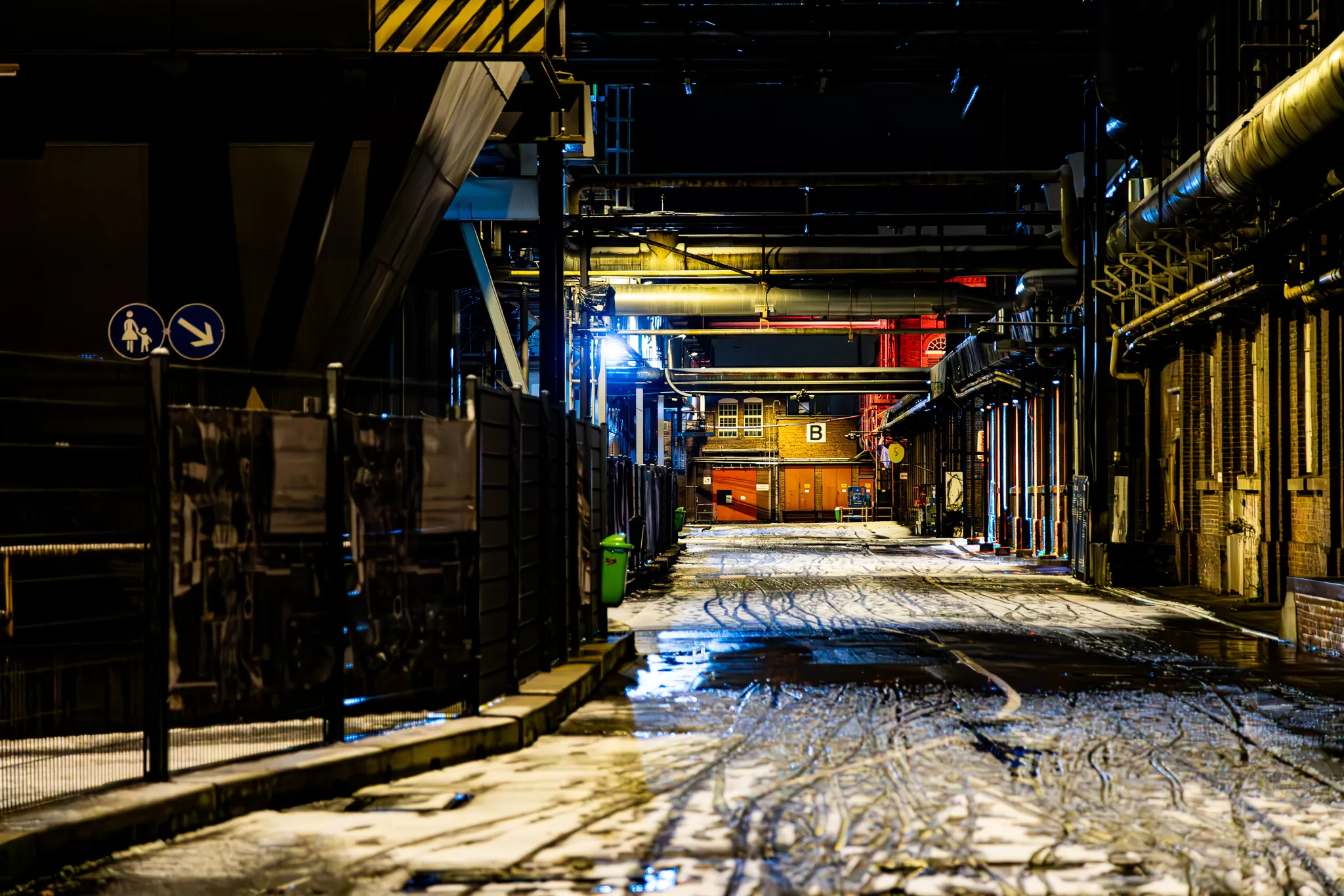 Werksgasse auf dem Zanders Gelände mit Fußgänger-Hinweisschild am Zaun, im Hintergrund ein rot beleuchtetes Tor mit der Aufschrift „B“, der schneebedeckte Boden spiegelt das Licht.
