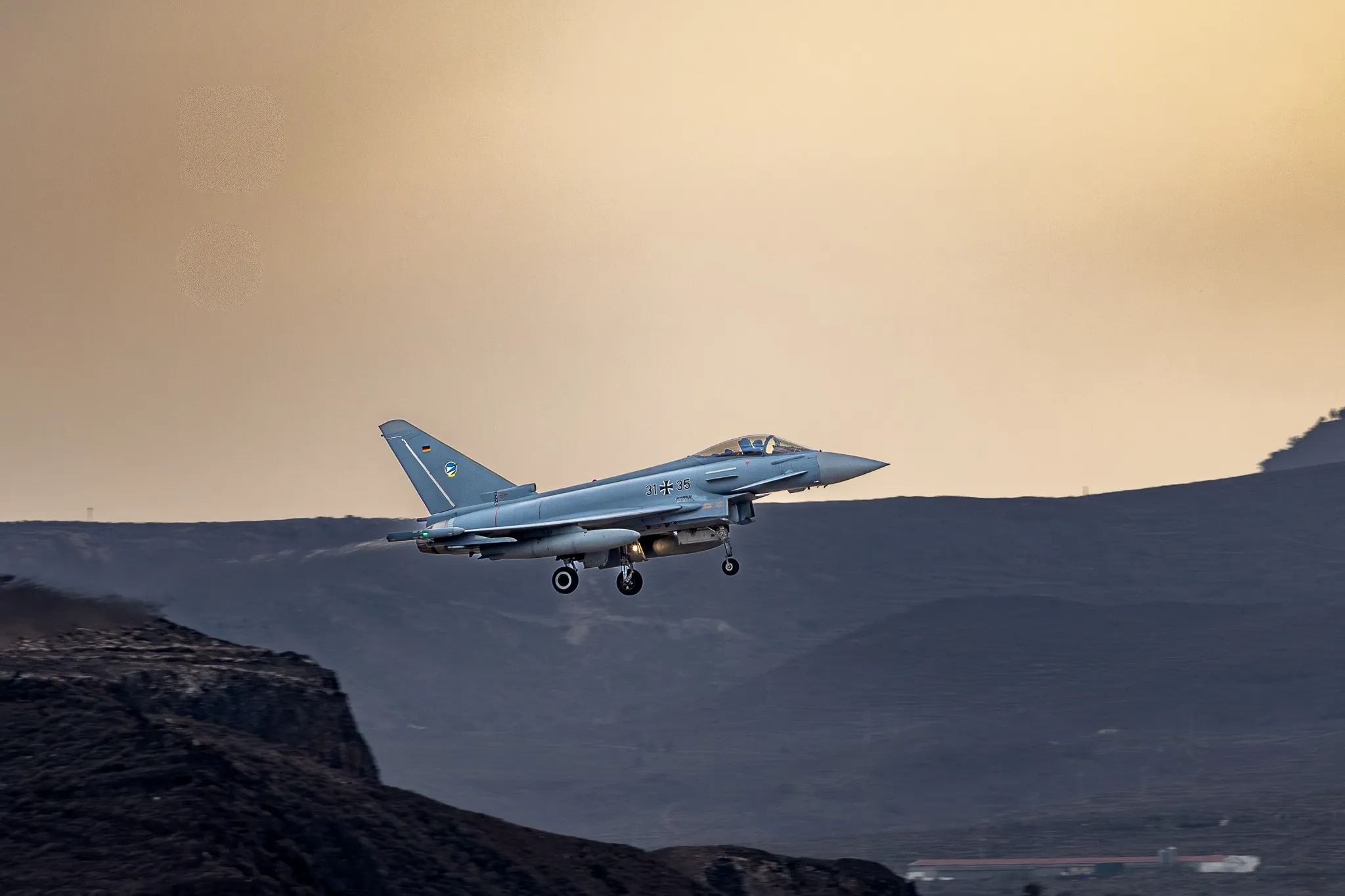 Ein Eurofighter Typhoon der deutschen Luftwaffe im Landeanflug mit ausgefahrenem Fahrwerk vor einer Bergsilhouette im warmen Abendlicht.