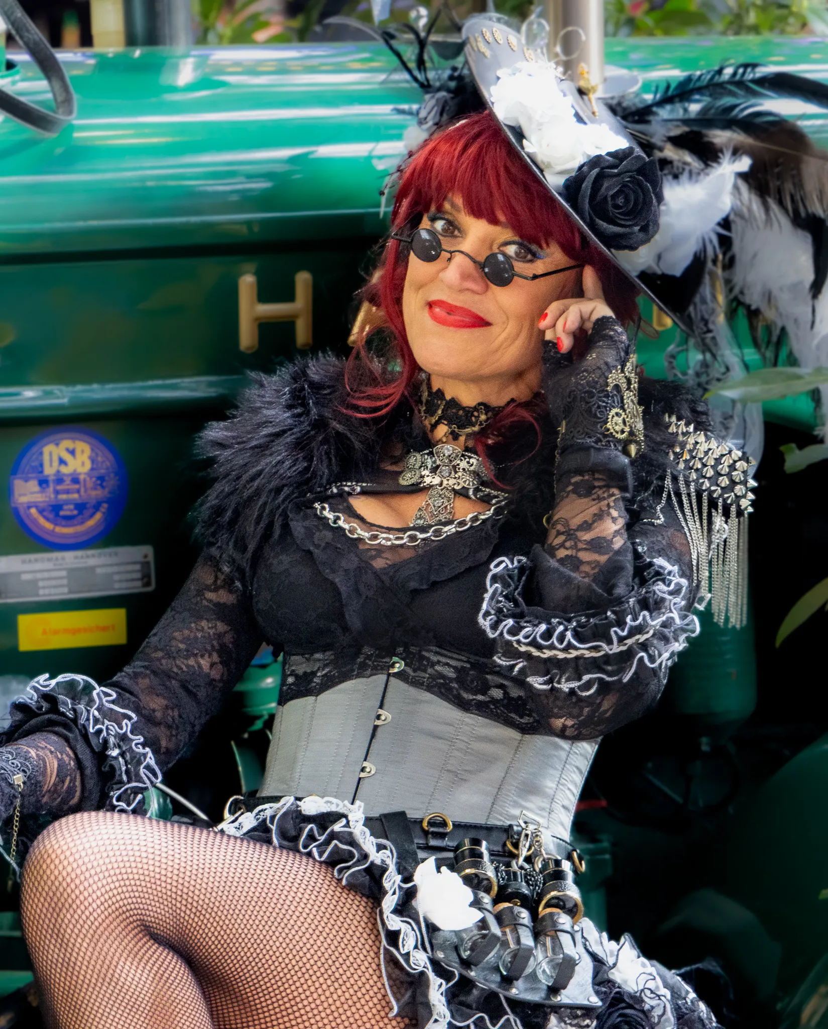 Frau mit rotem Haar in aufwendigem schwarzem Steampunk-Kostüm mit Pelzkragen, Netzstrümpfen und mechanischen Accessoires an einer grünen Zugmaschine lehnend.