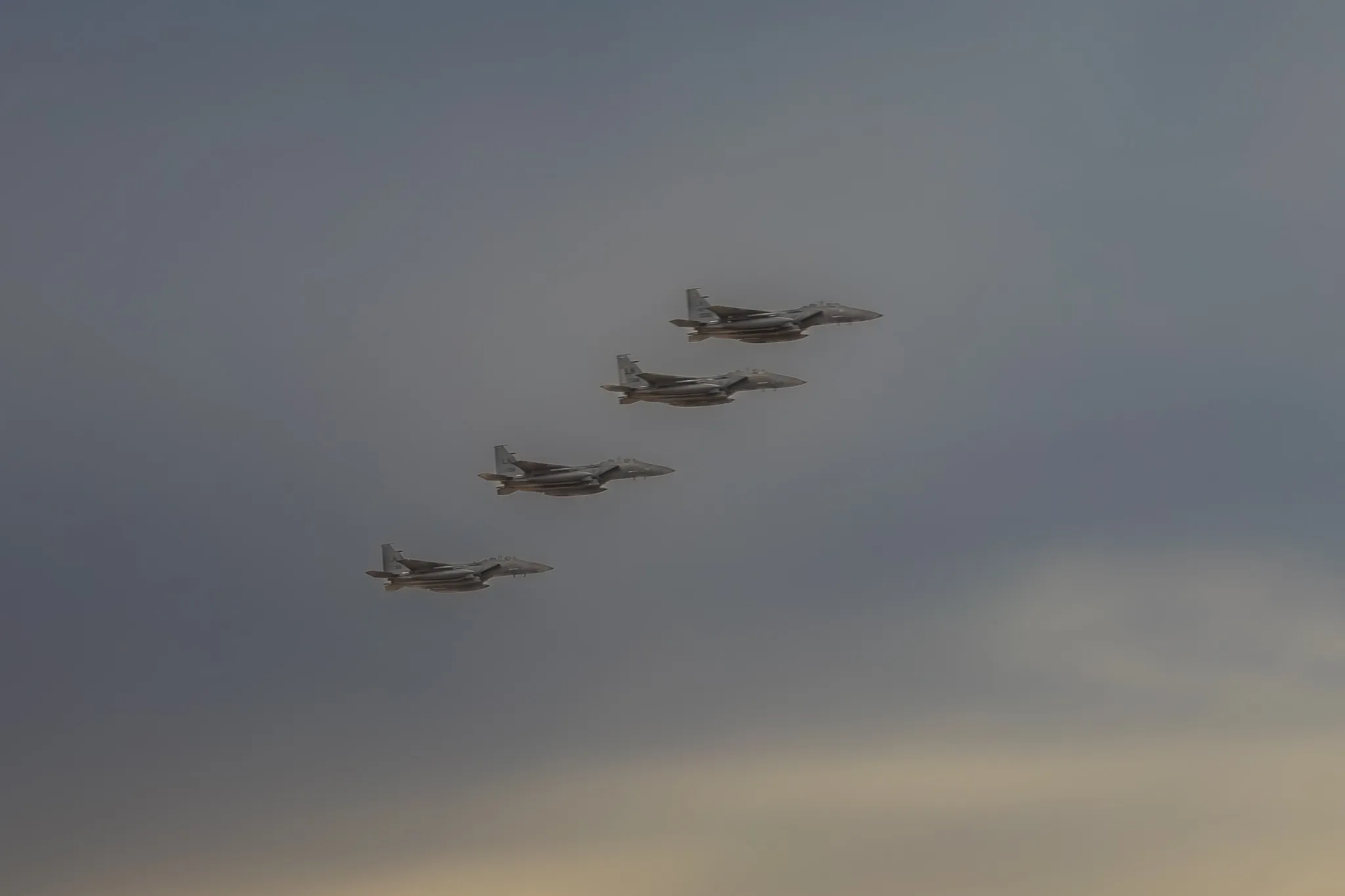 Vier F-15E Strike Eagle der US Air Force fliegen in gestaffelter Formation, Seitenansicht vor grau-diesigem Himmel.