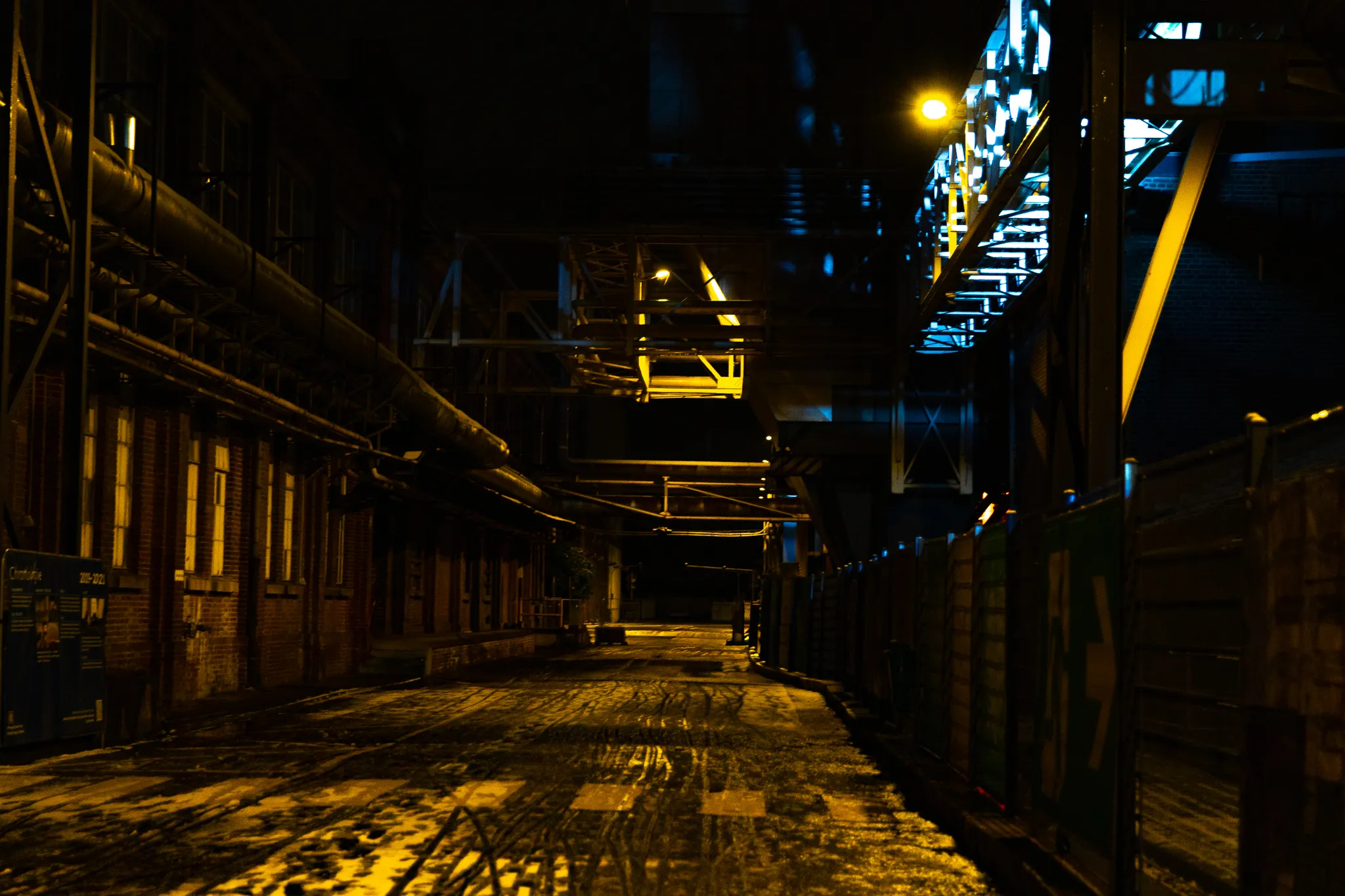 Nächtliche Gasse auf dem Zanders Gelände mit einer historischen Informationstafel links und einer bunt schimmernden Lichtinstallation an der Stahlkonstruktion rechts.