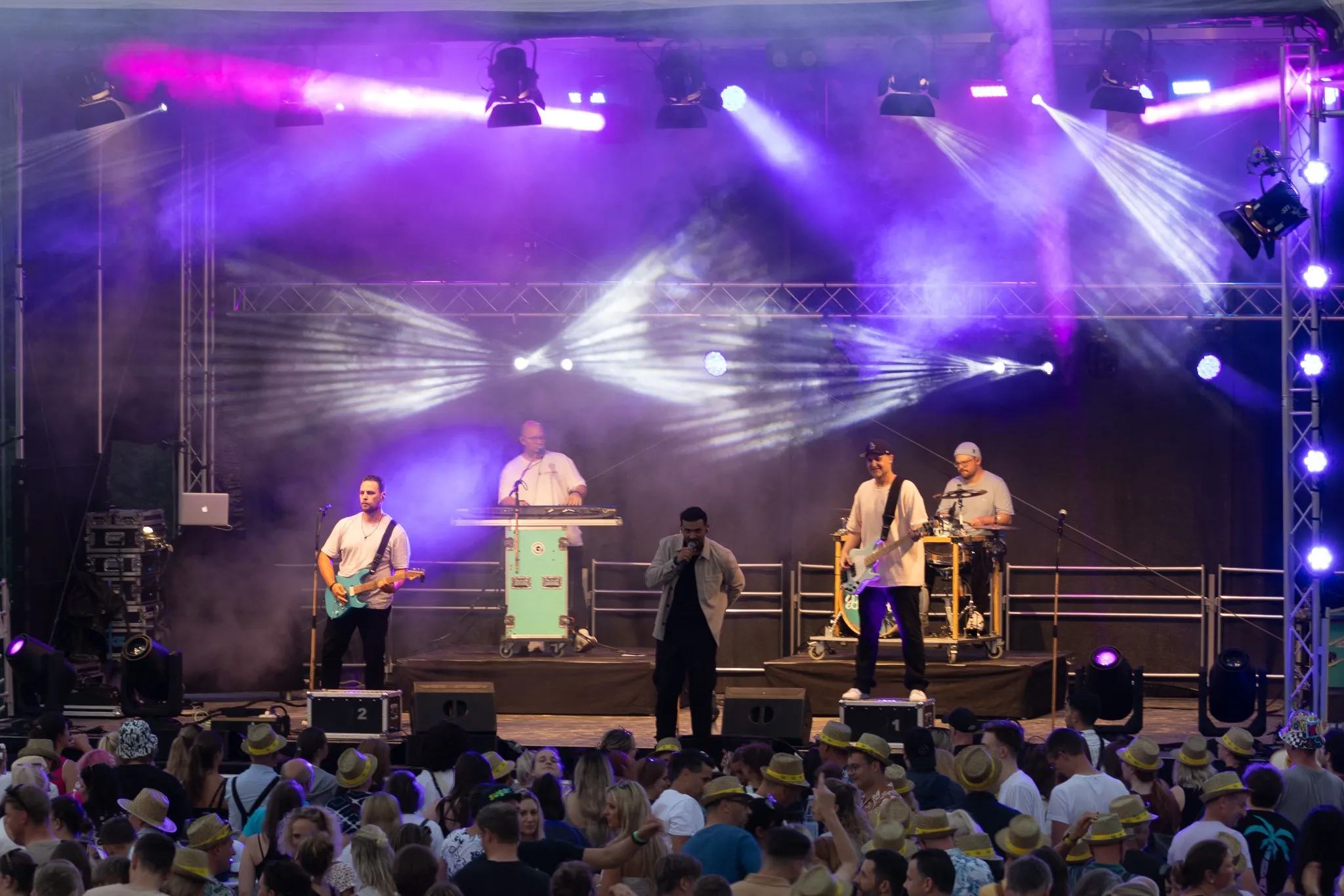 Band mit Musiker an türkisfarbener E-Gitarre, Keyboard-Setup und Sänger im hellen Jackett spielt vor dichtem Publikum, aus der Menge heraus fotografiert.