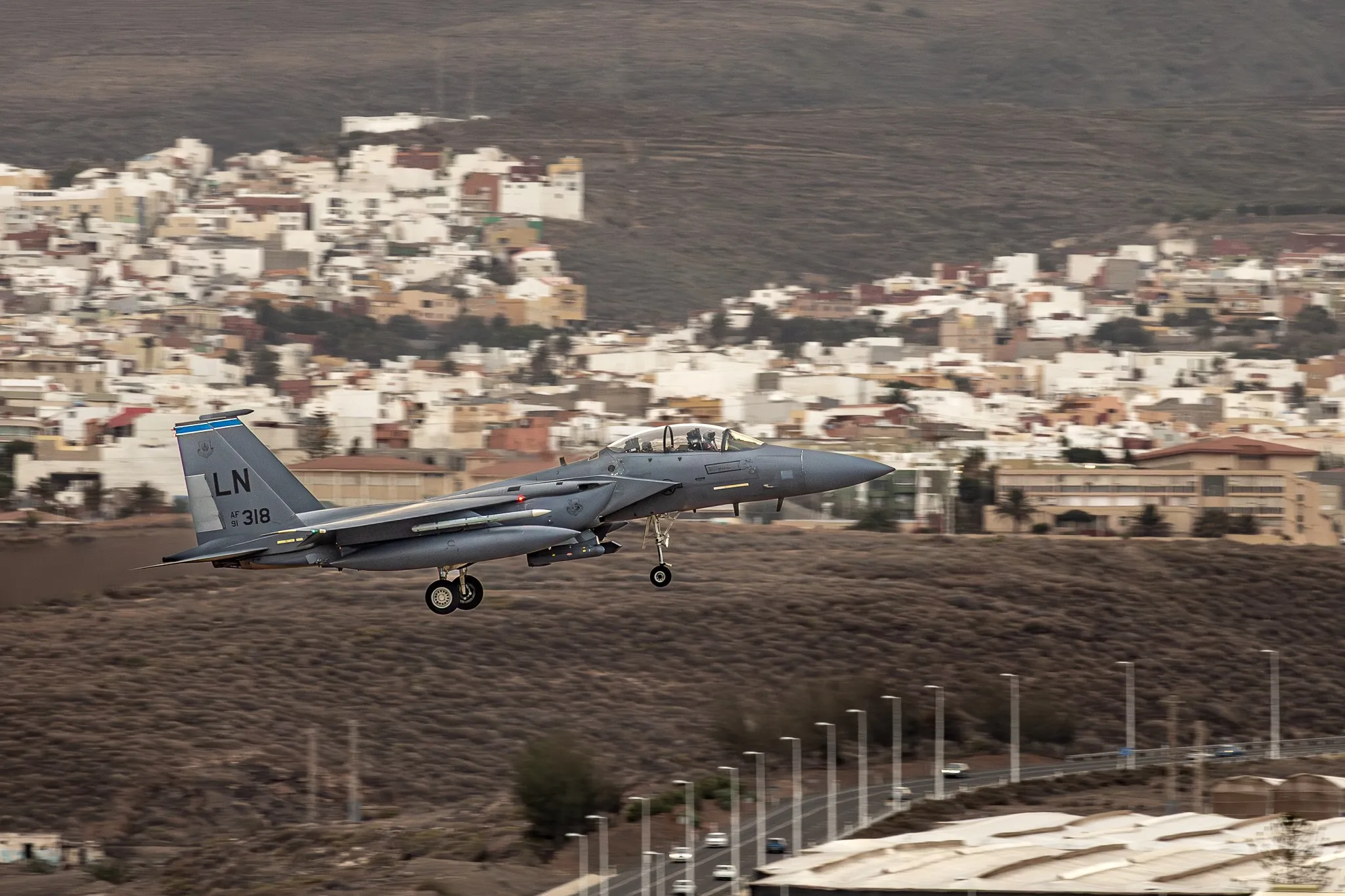 Eine F-15E Strike Eagle im Landeanflug mit ausgefahrenem Fahrwerk über einer Stadt am Fuß eines Berghangs.