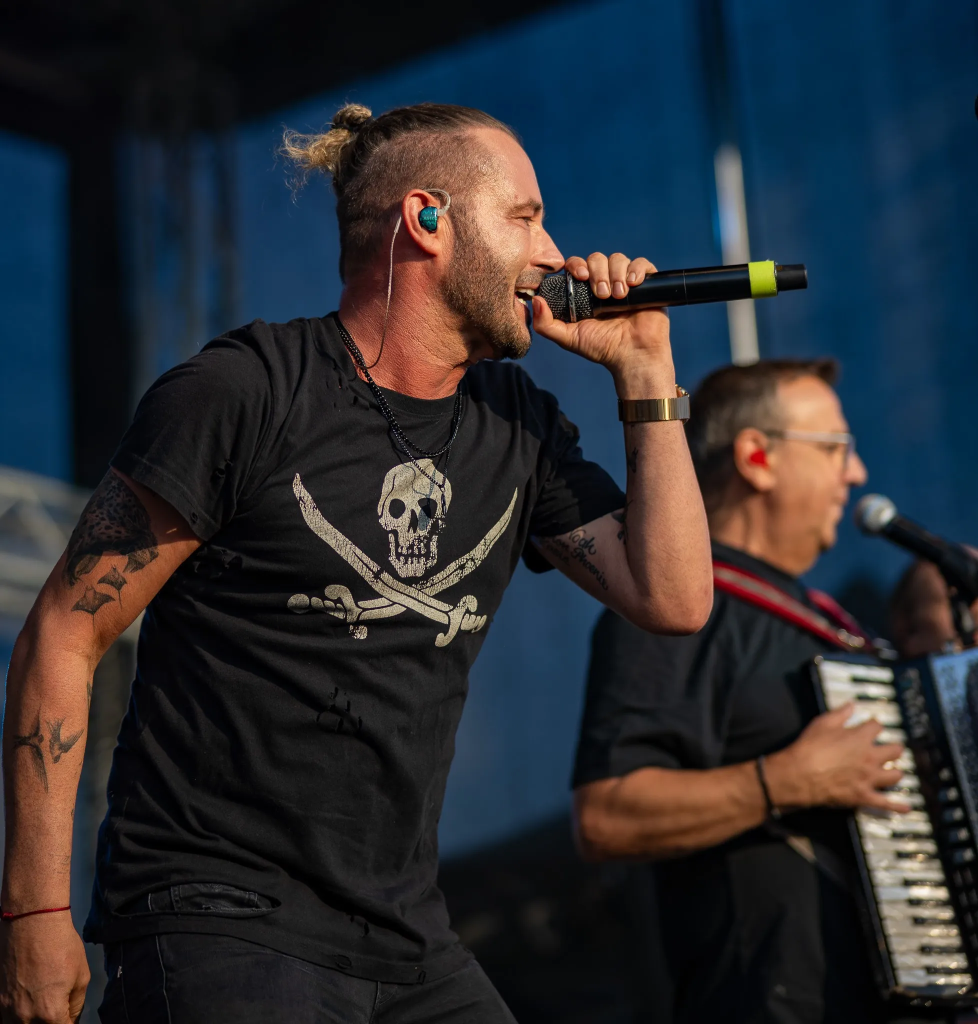Sänger mit Dutt und Tattoos singt intensiv ins Mikrofon, im Hintergrund unscharf ein Akkordeonspieler vor blauem Bühnenhintergrund.
