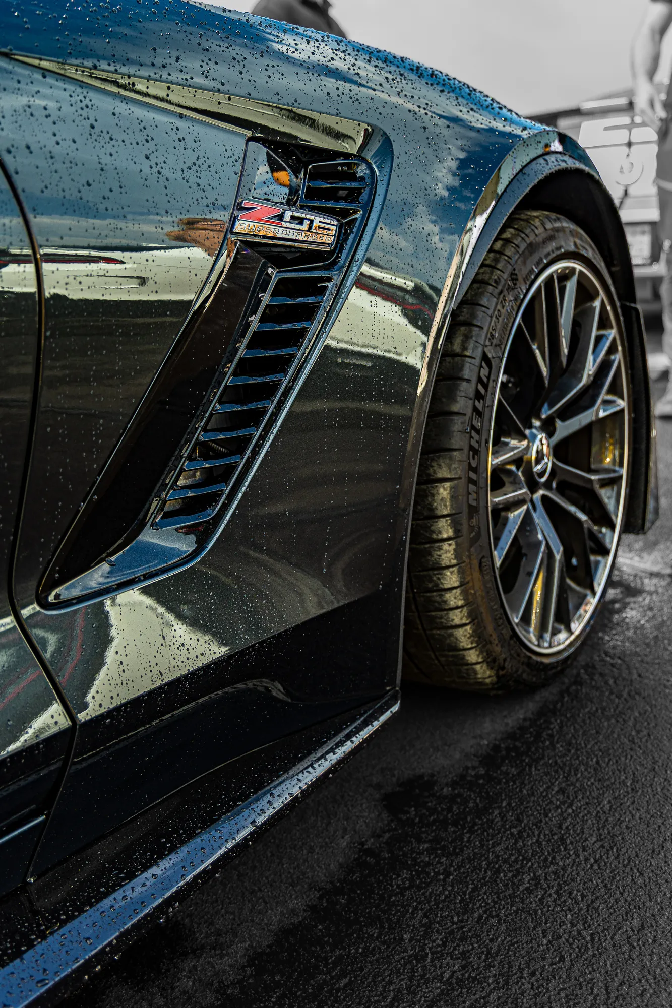 Nahaufnahme der hinteren Seitenpartie eines schwarzen Chevrolet Corvette Z06 Supercharged mit Lufteinlass-Emblem, verchromtem Rad und Michelin-Reifen, Regentropfen auf dem Lack.