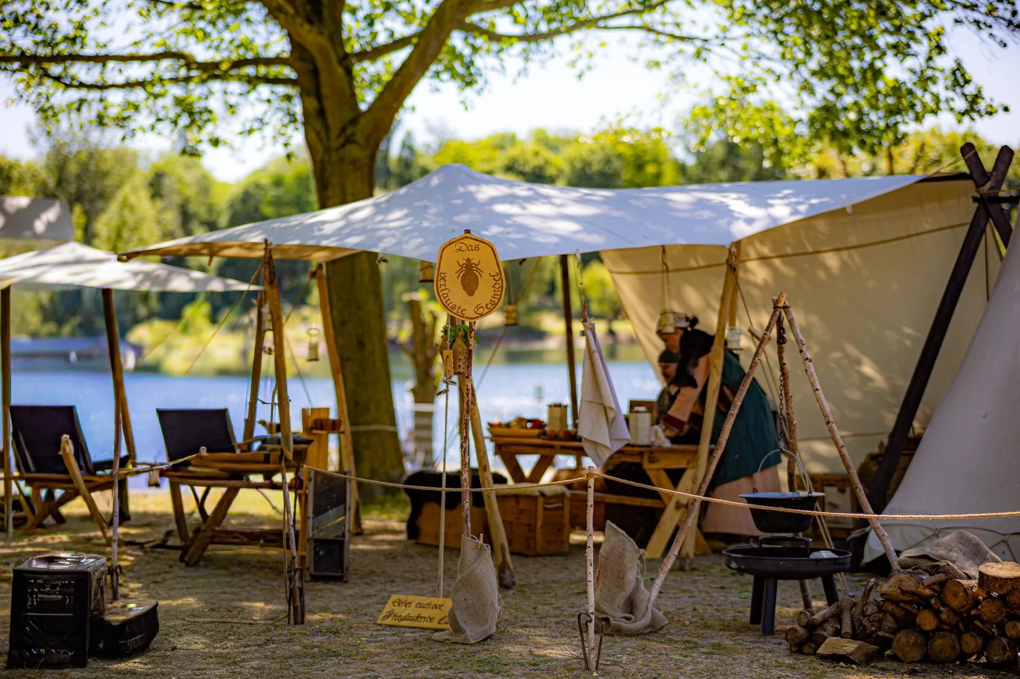 Lagerszene auf einem Mittelaltermarkt mit weißen Spitzzelten, Feuerstelle und Holzstapel, im Hintergrund Personen in historischer Kleidung an einem Tisch am Seeufer.