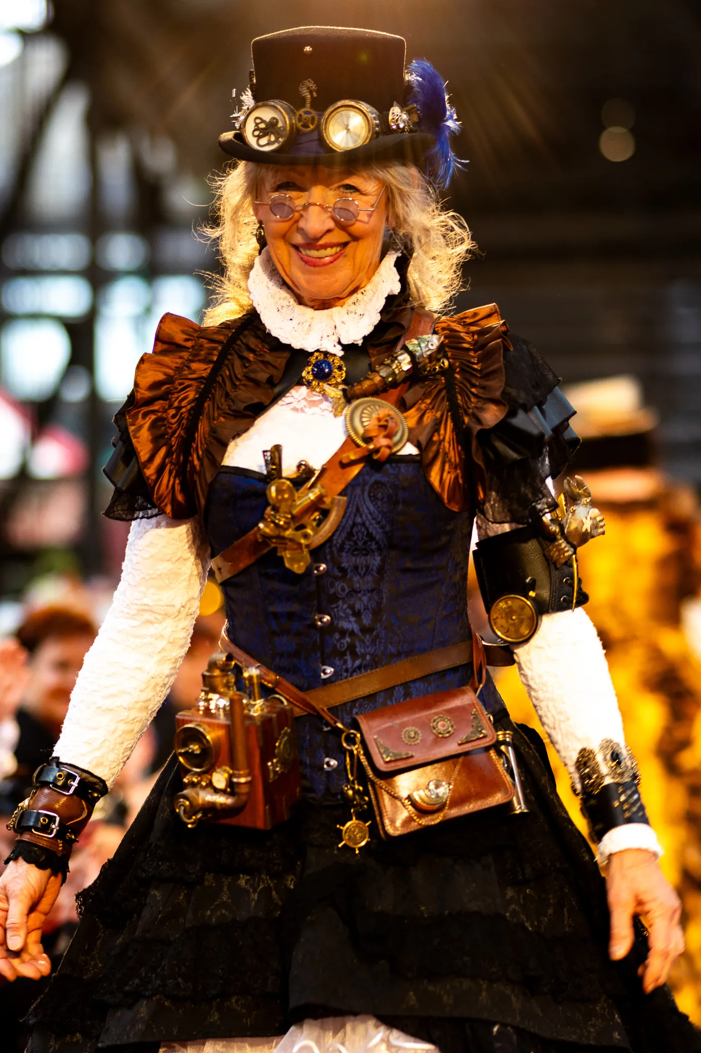 Lächelnde ältere Frau mit federgeschmücktem Steampunk-Zylinder, Rundbrille und aufwendigem Kostüm mit zahlreichen mechanischen Accessoires, Taschen und Gürtelapparaturen.