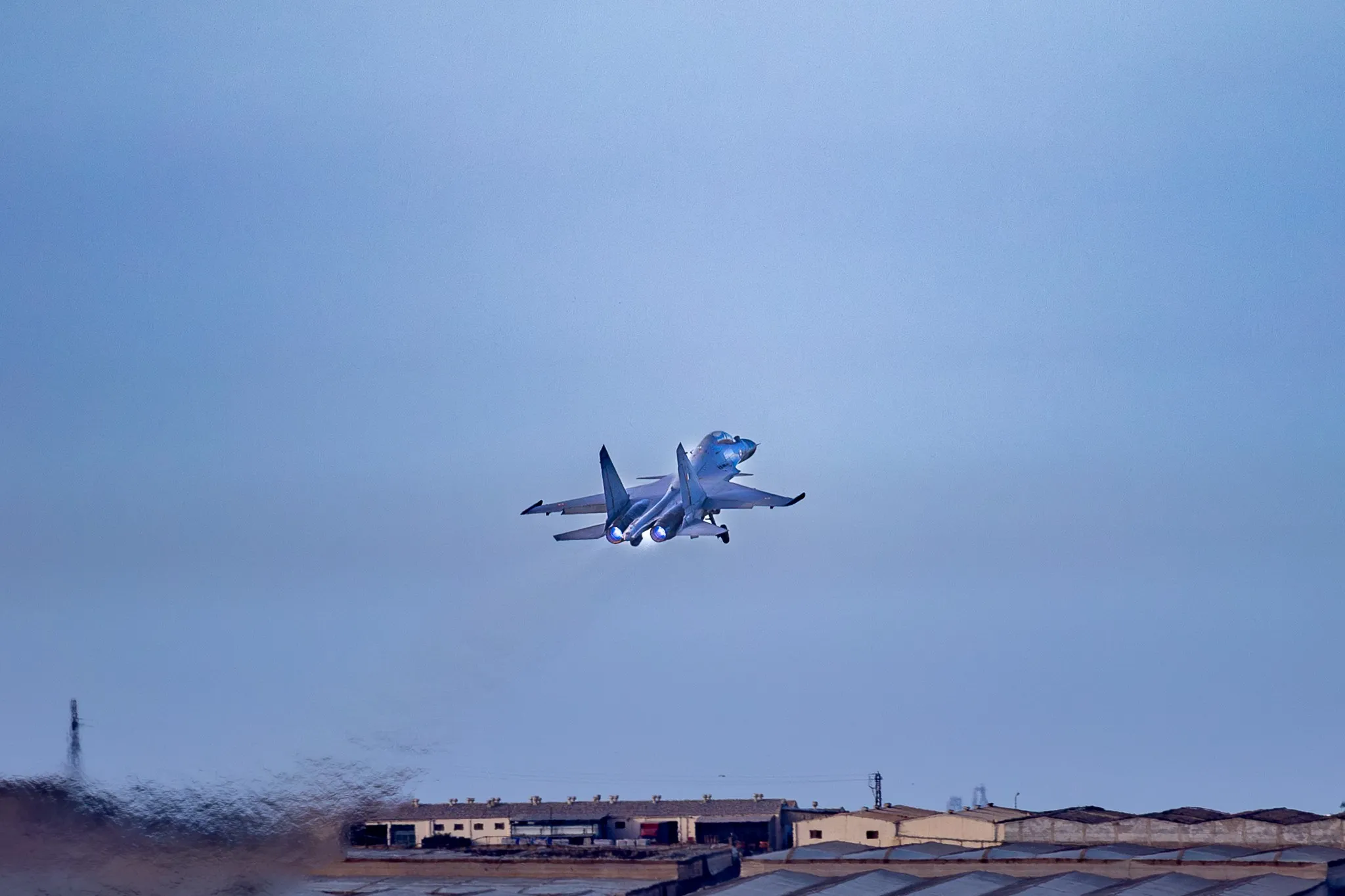 Ein Kampfjet mit deutlich sichtbarem, blau glühendem Doppel-Nachbrenner im steilen Steigflug kurz nach dem Start, Nahaufnahme vor blauem Himmel.