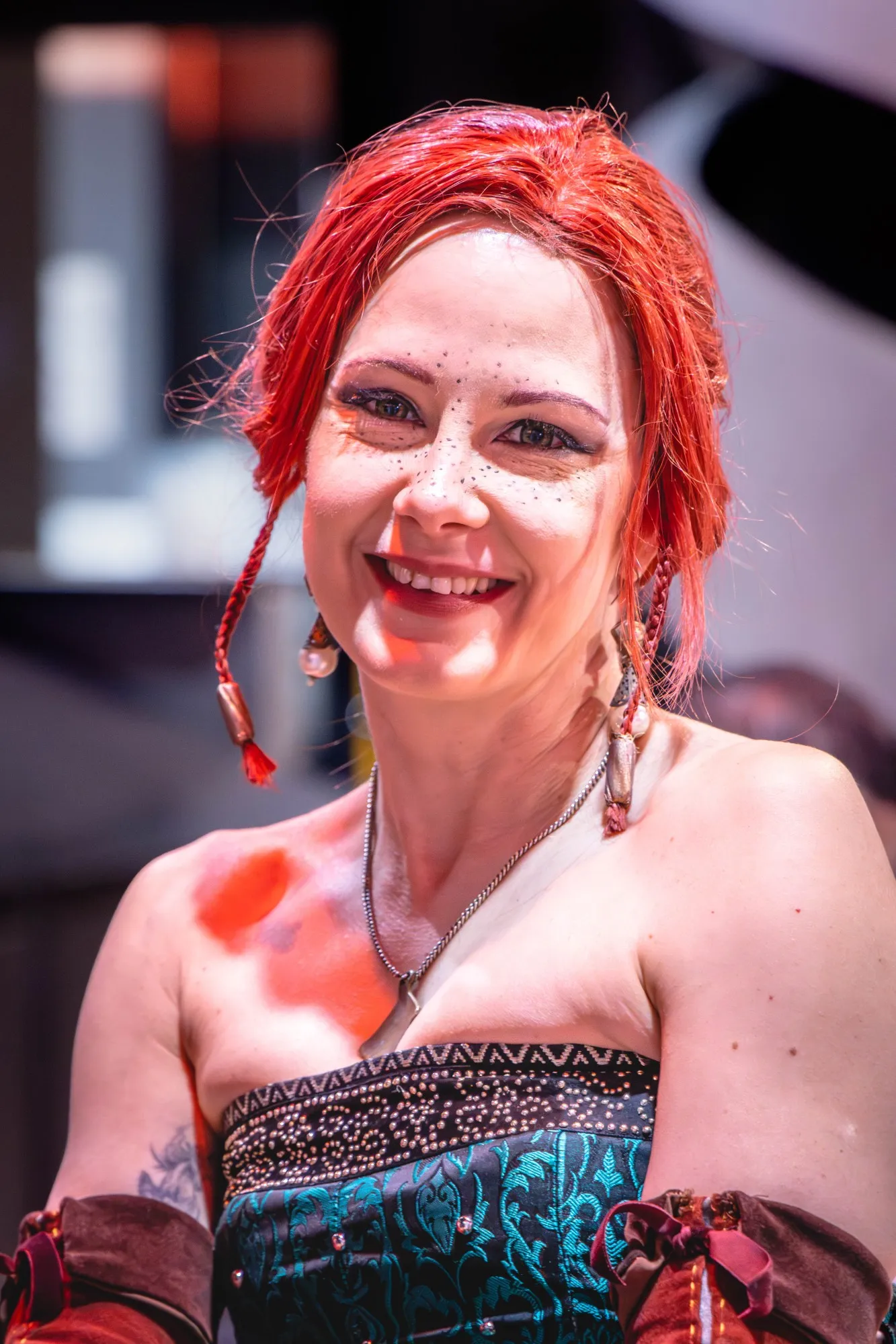 Lächelnde Cosplayerin mit leuchtend roten Haaren, aufgemalten Sommersprossen und geflochtenen Zöpfen, in schwarz-türkisfarbenem Mieder mit Nietenbesatz.