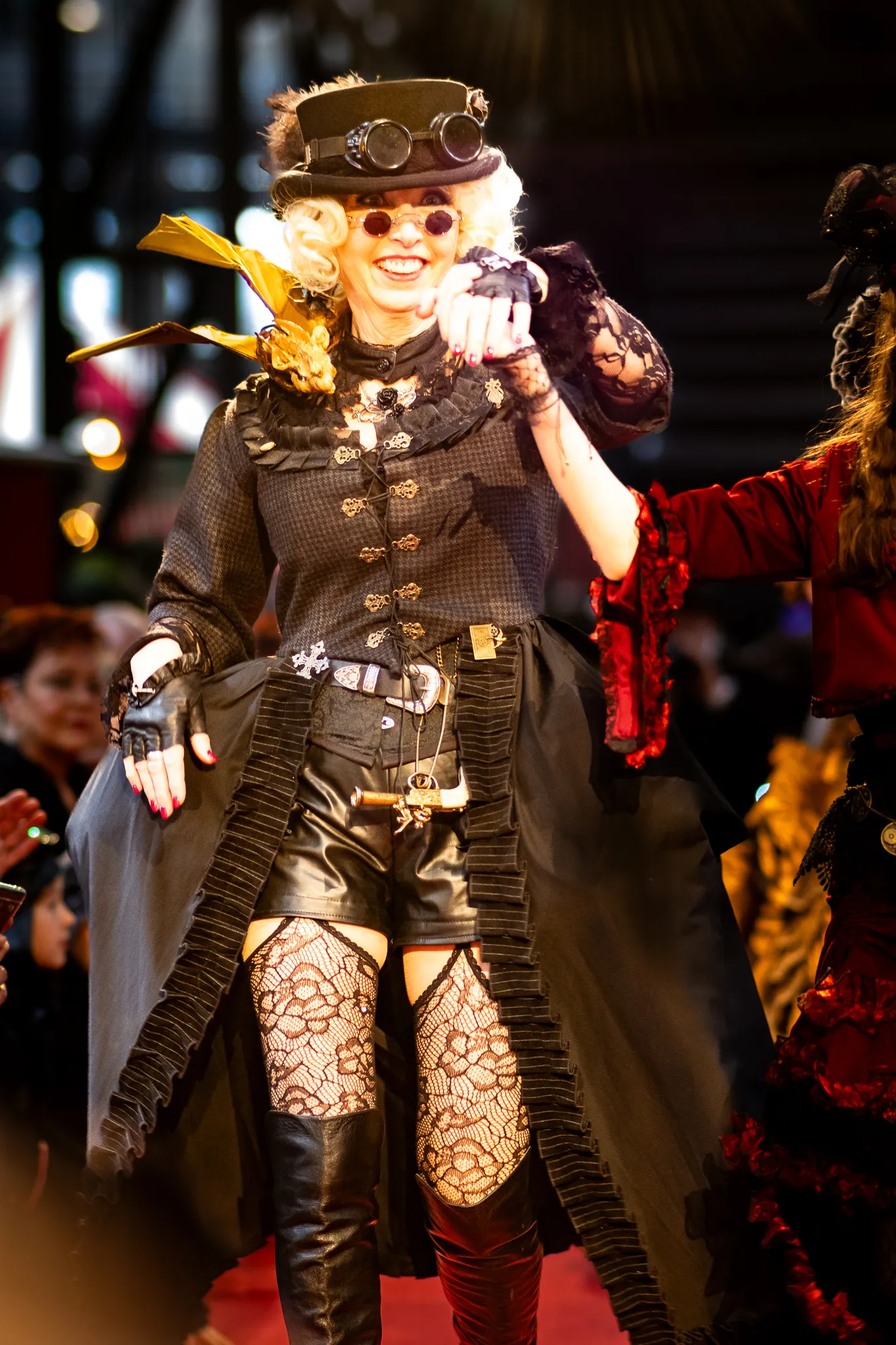Frau in schwarzem Steampunk-Outfit mit Fliegerbrille auf dem Hut, Netzstrümpfen und hohen Lederstiefeln, lachend und mit ausgestrecktem Arm in Bewegung, neben einer weiteren Person in Rot.