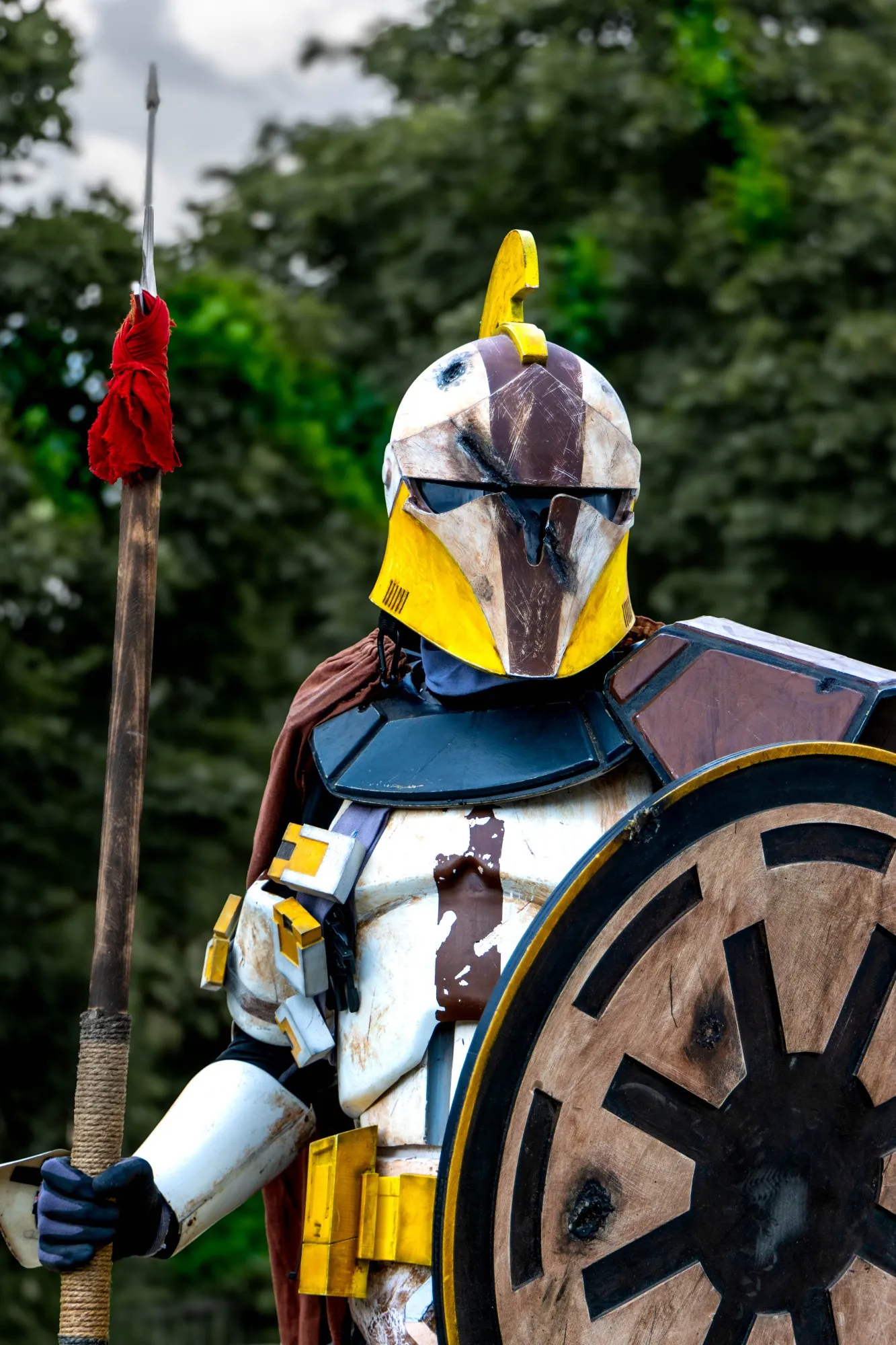Klontruppler-Cosplay mit verwittertem gelb-braun gemustertem Helm und roter Kapuze, hält Speer und Schild vor grünem Waldhintergrund.