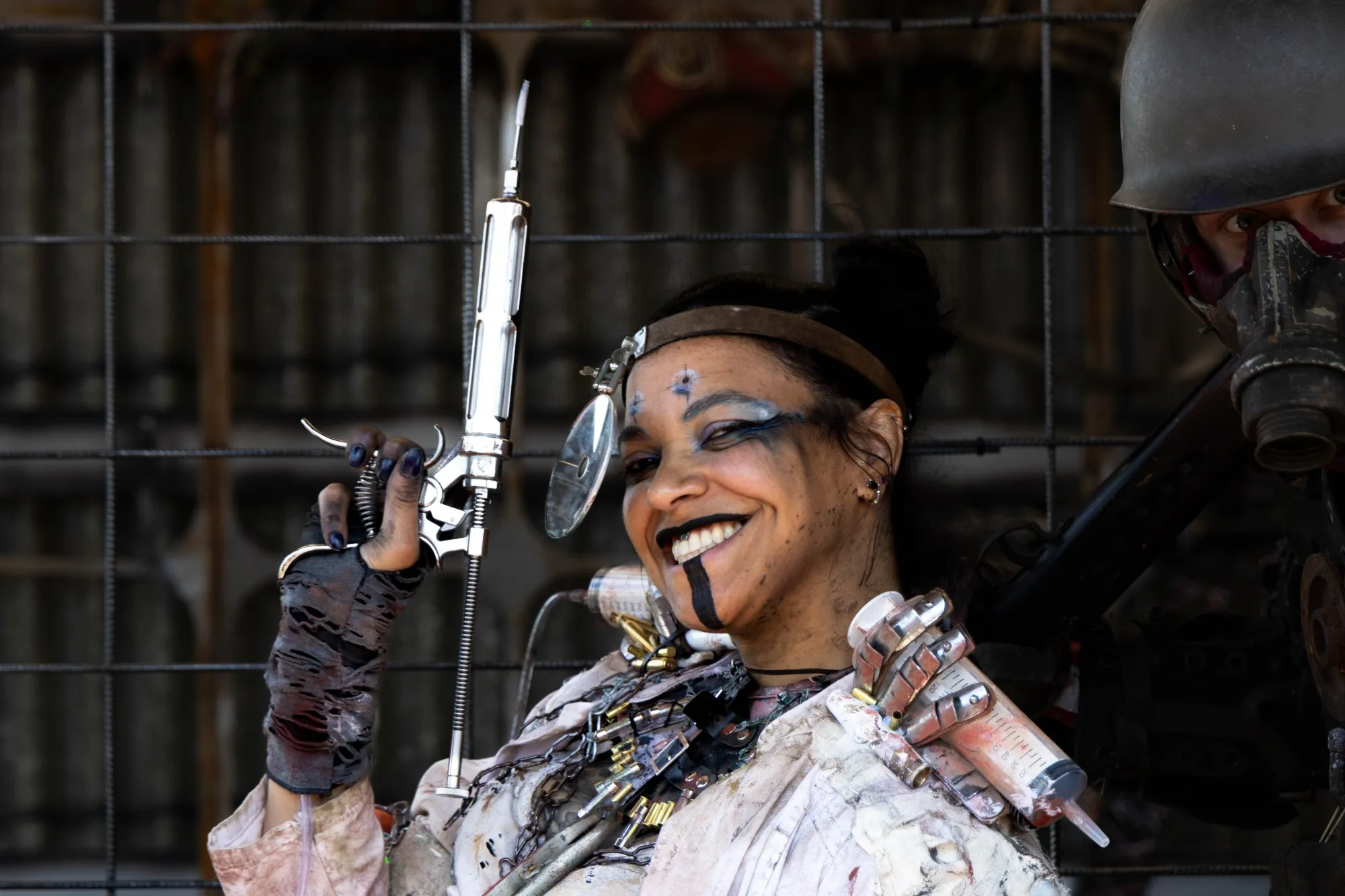 Lächelnde Cosplayerin in zerrissenem Endzeit-Outfit mit Munitionsgurten um den Hals, hält eine große metallische Spritze wie ein Werkzeug vor einem Metallgitter.