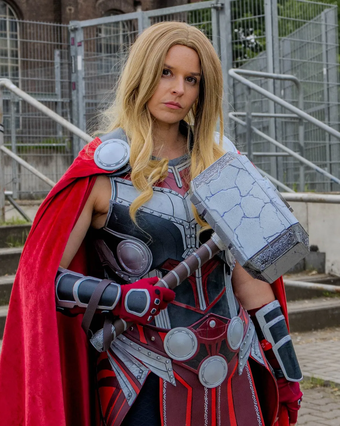 Cosplayerin als geflügelte Kriegerin mit blondem Haar, rotem Umhang und silbern-roter Plattenrüstung, hält einen großen Steinhammer vor städtischer Außenkulisse.