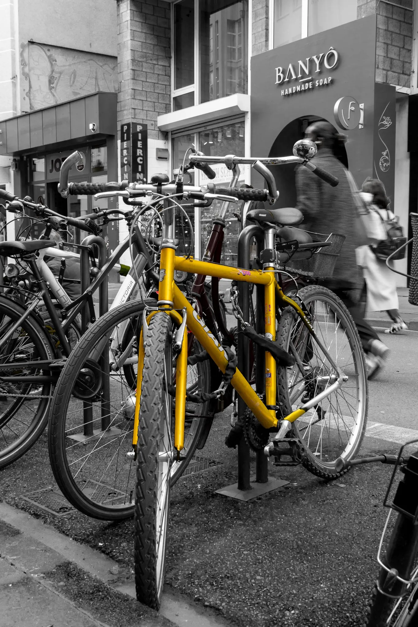 Gelbes Fahrrad an einem Fahrradständer, farblich hervorgehoben vor einer schwarz-weißen Einkaufsstraßen-Kulisse mit vorbeigehender, leicht verwischter Passantin.
